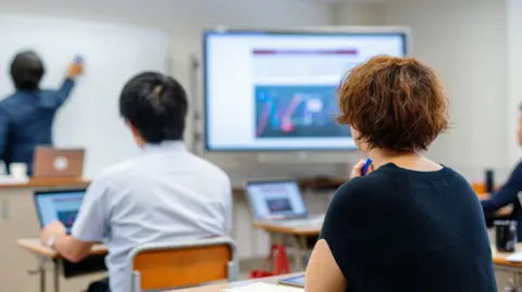 Getty Images Seorang wanita memegang pena di lehernya di ruang kelas saat dia melihat ke depan di mana seorang dosen menunjuk ke papan tulis 