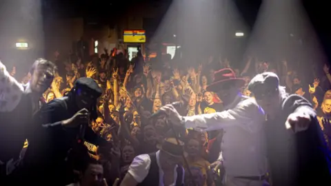 Music Venue Properties A crowd of people, lit in a yellow light all with their hands up. Everyone is posing to the camera. At the forefront of the image is four musicians, all posing for the camera.