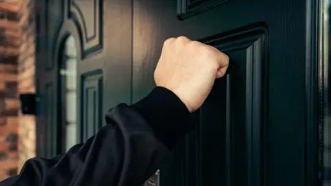 A hand knocking on a black door. The person is wearing a black long sleeve jumper.