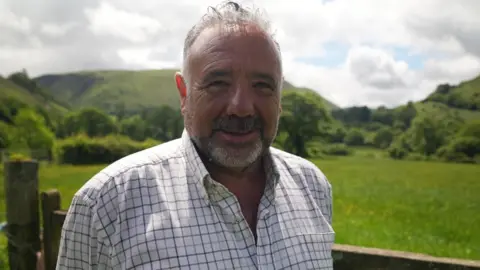 A man smiling at the camera. He has short grey hair, a grey beard and is wearing a white and black check shirt