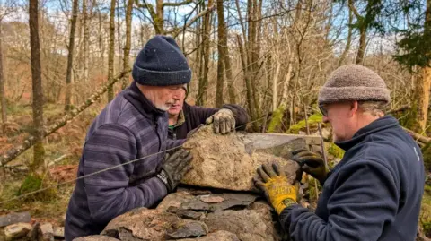 Three men lift a stone on to a wall. They are all wearing fleeces, wooly hats and gloves. A number of trees can be seen behind the wall.