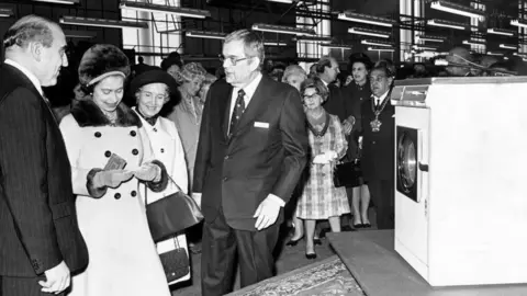 Getty Images Two men wearing dark suits, looking at a woman in a fur-lined coat and fur hat. In front of them is a washing machine, and behind them a crowd of onlookers. Above them are factory strip lights.