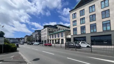 BBC A series glass and limestone finance buildings, next to a wide road, leading up to a roundabout.