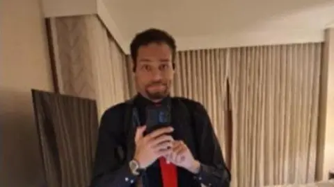 A man wearing a black button-down shirt and red tie holds a phone as he takes a photo of himself