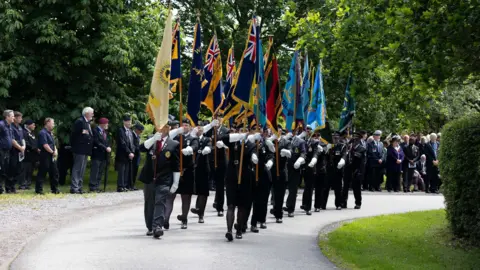 Bristol Memorial Woodlands A parade is making its way through Bristol Memorial Woodlands, with different British Army flags being carried. There are crowds of onlookers including veterans and army personnel.