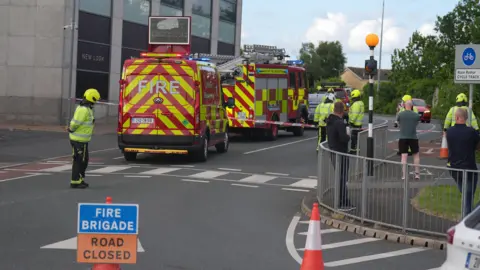 PA Media Three red and yellow fire service vehicles sits in a line outside a grey bricked building. They have cordoned off the area using red and white tape. Four fire men in illuminous yellow jackets can be seen beyond the cordon with one in front of it. A gathering of three men watch on from in-front of the cordon. A blue sign in white font reads 'Fire Brigade'. An orange sign in black font reads 'Road Closed.'
