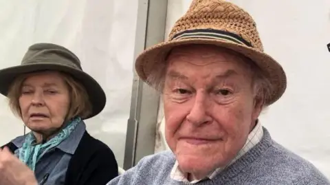 Martin Heath/BBC A close-up selfie-style photo of two people seated side by side in an outdoor event space. In the centre, Timothy West is sitting slightly behind the camera holder, wearing a woven brown hat and a light grey sweater over a collared shirt. On the left, Prunella Scales is partially in frame, seated further back. She is wearing a darker hat, a scarf, and a jacket, and is positioned slightly turned away from the camera.