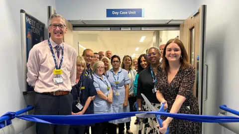 UHNM Dr Simon Constable and MP Leigh Ingham, who is holding a large set of scissors, are stood behind a blue ribbon. There are a set of doors behind them and nurses in uniform.