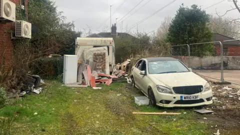 Photograph of flytipping on Baguley Crescent in Prestwich. The image shows Bags of waste, furniture, mattresses and an abandoned car. The Mercedes has been registered as off the road (SORN). The MOT expired on 9th February 2024.