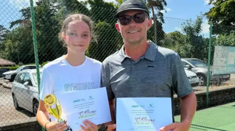 Steve Daniel A man and his teenage daughter pose and smile for the camera. They are both holding certificates while the girl is also holding a gold trophy. The girl wears a white T-shirt and has brown hair that is tied back. The man wears a black cap, sunglasses and a grey polo shirt. 
