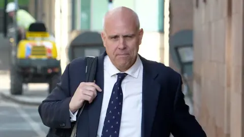 Richard Wright KC wearing a navy suit and tie walking down the street towards Newcastle Crown Court with a bag over his shoulder.