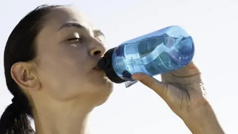 A young woman is cooling down drinking a bottle of water. She has long, dark hair which is tied back