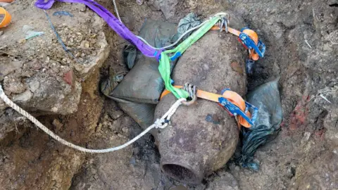 Royal Navy Straps have been fastened around the bomb which is sitting in the mud. Sandbags have been placed around the bomb.