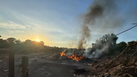 A fire and black ash left over from a bonfire. Smoke is rising. It's morning. Trees are in the background.