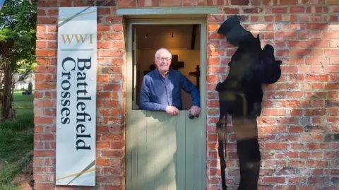 Cheltenham Civic Society A man with white hair and glasses standing behind a stable door which is closed on the bottom. He is standing in a small brick outhouse building with a vertical sign on the left that says 'World War One battlefield crosses', with a silhouette of a soldier on the right of the door.