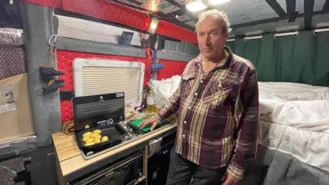 Dave Knight is standing inside his caravan cooking potatoes. A bed is right behind him and he is wearing a checked shirt.