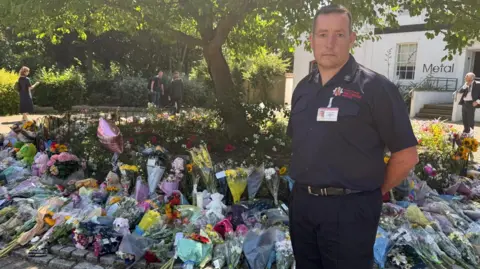 A man standing in his uniform with a background of flowers spread across a tree. Teddy bears are also being left. The man has a sombre expression on his face and he has his hands behind his back.