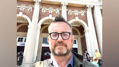 Matthew Carr Matthew Carr stands outside Clacton Town Hall. He is wearing a grey overcoat, and blue corduroy shirt with a bag strap slung over his shoulder. He has a tuft of hair at the front and is wearing black-framed glasses. He has a grey/brown beard.