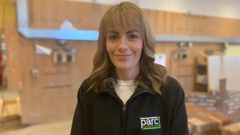 A woman with long blonde-hair and a black zip top with a Parc Construction logo on it stands in a bricklaying workshop