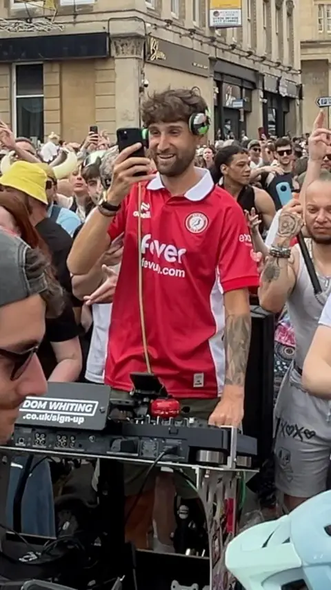 DJ Dom Whiting, wearing a red Bristol City top, on his bike with DJ equipment in front of him. Behind is a large crowd of people.