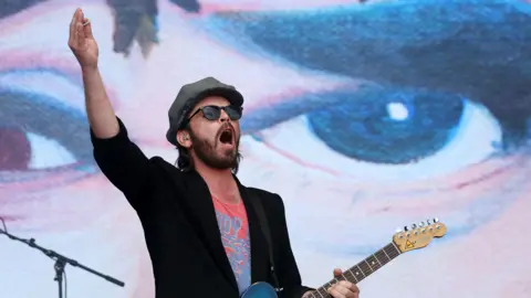 EPA Gaz Coombes wearing a black jacket and grey hat, holding a guitar and putting his other arm in the air.