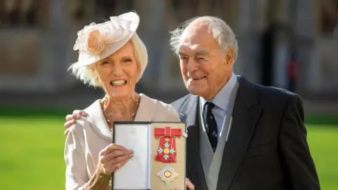 Getty Images Berry with Hunnings at Buckingham Palace holding her award. She is smiling and wears a light pink dress, a pink hat and a gold necklace. Paul, on the right, wears a navy suit and grey waistcoat.