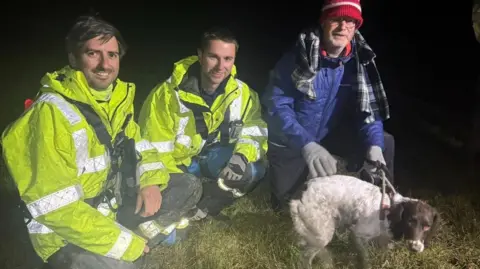 Willow, a brown and white spaniel, beside three men crouching down on grass in the dark, two in high-vis yellow jackets and the other in a blue hat and red woolen hat.