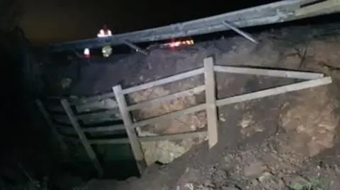 Bent crash barriers along the top of a bank of earth, with a broken wooden fence on the side of the bank. Traffic headlights appear in the background.