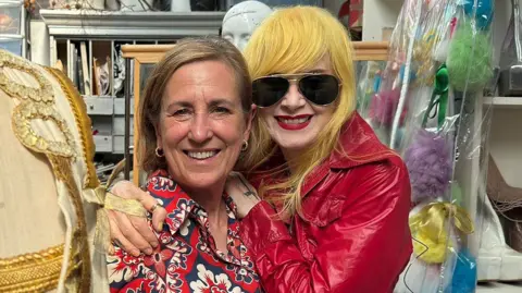 Kirsty Wark, in a red tile-patterned dress, is hugged by red PVC dress-wearing designer Pam Hogg, who is wearing striking yellow hair and sunglasses. A beige and sparkly military-style outfit is on a tailor's dummy in the foreground, a storeroom behind the pair, with shelves displaying platform shoes and many bags, boxes and drawers around.