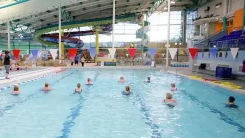 IOM GOVERNMENT People lined up in the main pool at the NSC. It has lane markings on the bottom and swimming bunting hanging above it. Water slides can be seen at the other end.