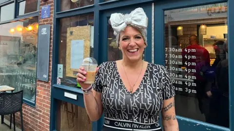 BBC/Richard Fox A woman holds up an iced coffee outside a coffee shop. She wears a headscarf with a silver bow tied at the front, and a black top with the Calvin Klein logo repeated in a pattern across it. 