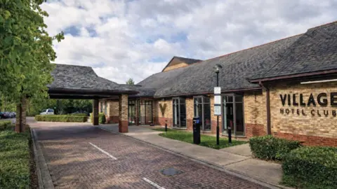 VUR Village Properties Ltd / Broxbourne Borough Council A computer-generated image of the front of a hotel. It has one storey with the words "Village Hotel Club" written on the side. There is a covered porch at the front over a driveway which leads to a car park in the background.