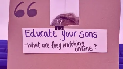 This Life Collective A cardboard sign is held in front of a girls face, her hands are visible on the edges. 'Educate your sons - what are they watching online?' is written in black marker pen. 