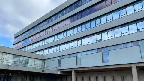 A view of a brutalist multi-storey 1960s building clad in grey, with windows stretching the full length. The picture is taken from the ground outside, where stairs lead up to a covered walkway into the building. 