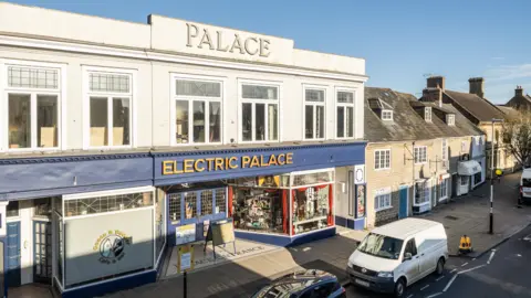 Symonds and Sampson Elevated view of the exterior of Electric Palace. The frontage is two storeys with a tiled recessed entrance flanked by two shops. Above the entrance is a blue sign with Electric Palace mounted in gold letters. Above the first floor windows is a white parapet with the word Palace mounted in large letters.