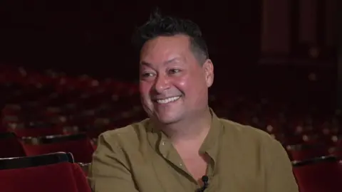 A man with short black hair smiles while sitting in theatre stalls. He is wearing a green shirt