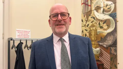 BBC Professor Andrew Le Sueur is smiling at the camera. To his left is a painting showing a chalice and the Jersey flag and behind him is a coat rack. He is wearing glasses and a pink shirt, patterned tie and navy jacket. 