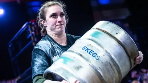 A woman during a strongwoman competition carries a large metal keg. She has dark hair that has been plaited back and wears a black T-shirt with chalk over it.