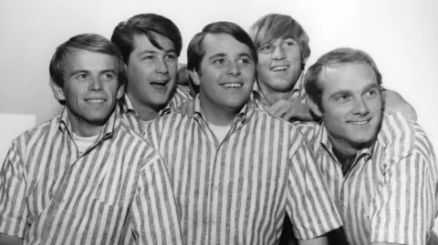 The five members of The Beach Boys pose for a photo. They're all wearing identical stripes shirts. The picture is in black and white.