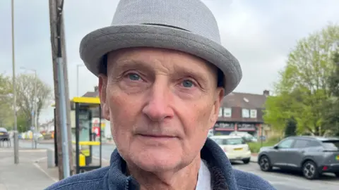 Alan stands for the photo, he stands on a pavement. He's wearing a grey hat and has blue eyes. He is an older man.