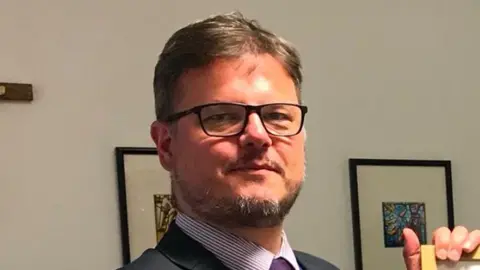 St Robert of Newminster School Dean Juric has glasses and a beard, his hair is brown and grey. He is wearing a dark suit with a purple striped shirt and purple tie. In the background is a crucifix depicting Jesus and he is holding a framed Duke of Edinburgh Award certificate, which is out of frame.