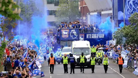 PA Media Police officers escort the large blue bus away from Portman Road stadium. Large crowds of people are gathered either side of the road and several blue flares have been lit.