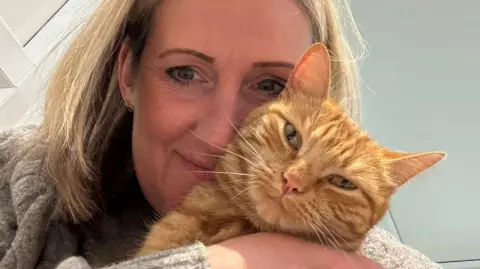 Hedgerows Cattery A woman with blonde hair wearing a beige jumper holds a ginger cat