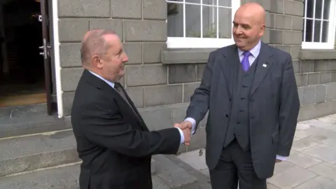 Two men shaking hands outside the States of Guernsey's Royal Court - a grey brick building. On the left is Deputy Allister Langlois, a small man with thinning grey hair wearing a black suit and a checked tie. On the right is Deputy Jonathan Le Tocq, a bald white man with a navy blue suit on, a waistcoat, purple shirt and purple tie. 