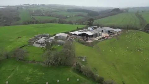 Smeaton Farm near Saltash is seen from above, with various barns and outbuildings seen scattered across an area surrounded by green fields and hedgerows. 