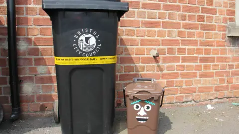 Bristol Waste A black bin with the Bristol City Council logo on it. Wrapped around the bin is yellow plastic tape which reads 'Slim my waste, No food waste'. Next to it is a small brown food waste bin with cartoon face stickers on it.