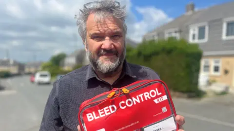 A man with grey hair and a beard looking at the camera, he is wearing a dark grey shirt and has glasses pushed back on his head. There is a street with houses in the background and he is holding a red case which says bleed control kit in white lettering