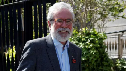 PA Media Gerry Adams wears a grey suit jacket with two pins on his lapel, a light coloured shirt with the top button undone and a pair of circular brown glasses. He is smiling at the camera as he walks past, where the gates of the High Court, as well as some trees, are behind him. Mr Adams has white hair and a beard.