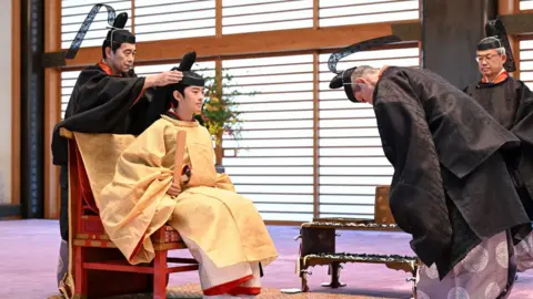 A man in ceremonial dress attaches a ceremonial hat to a young man.