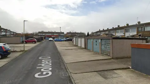 Google maps Golden Close in Tuffley, which is a residential area, during the day time. There are 7 garages to the right with a row of terraced houses behind them. 